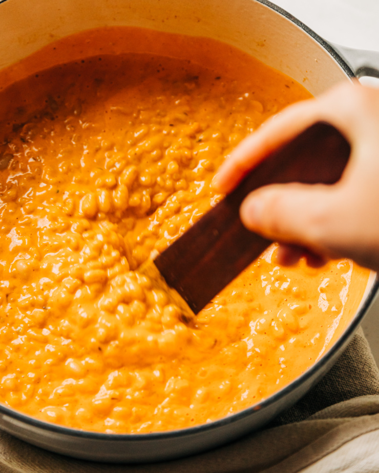 A slight 3/4 angle image shows a hand using a wooden utensil to stir up a "cheesy" vegan barley and white bean base.