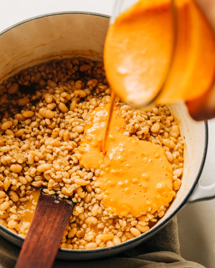 An overhead shot shows a creamy vegan red pepper and cashew sauce being poured into a pot of cooked barley and white beans.
