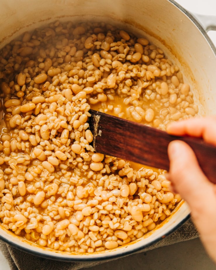 An up close, overhead image shows a hand using a wooden tool to stir up a pot of cooked white beans and barley.