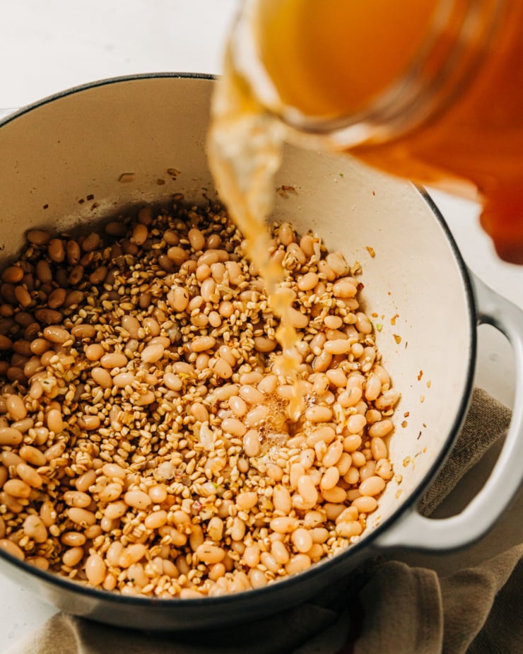An overhead shot shows vegetable stock being poured into a pot with white beans and barley.