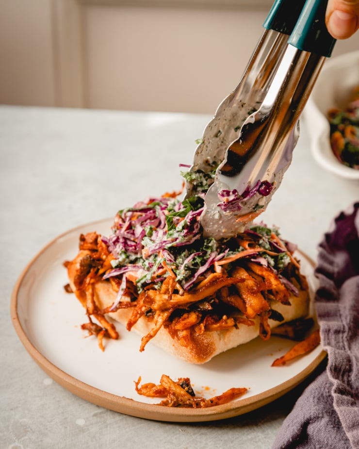 A slight 3/4 angle shot of tongs dropping some creamy slaw on top of BBQ pulled mushrooms resting in a bottom bun.