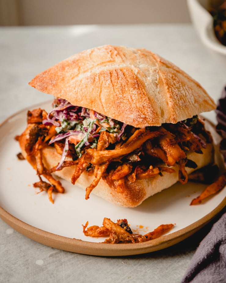 A slight 3/4 angle image of a crispy BBQ pulled mushroom sandwich topped with slaw on a small white plate.