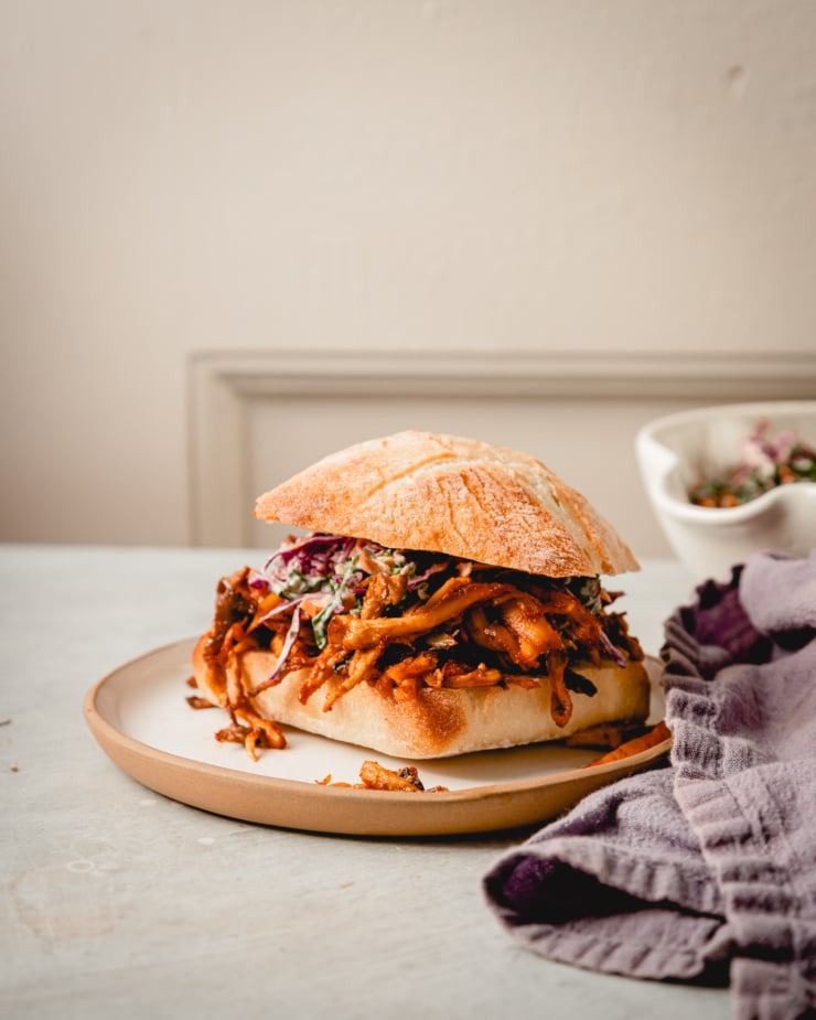 A head on shot of a crispy BBQ pulled mushroom sandwich with slaw on top. A linen napkin is nearby.