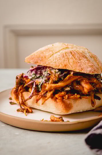 An up close, head on shot of a crispy BBQ pulled mushroom sandwich with slaw on top. A linen napkin is nearby.