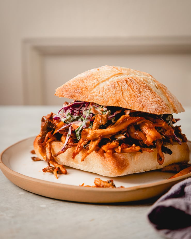 An up close, head on shot of a crispy BBQ pulled mushroom sandwich with slaw on top. A linen napkin is nearby.