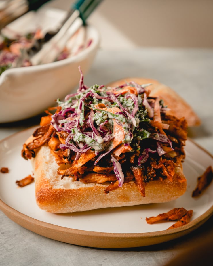 A slight 3/4 angle of some kale and red cabbage slaw on top of a mound of BBQ pulled mushrooms on the bottom half of a ciabatta bun.