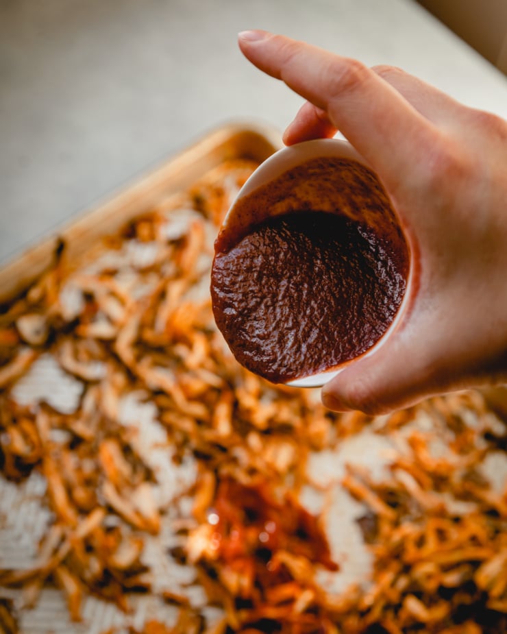 An overhead shot shows a hand pouring a bowl of BBQ sauce over some roasted mushrooms.