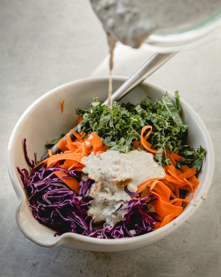 A slight 3/4 angle shows tahini ranch dressing being poured onto shredded red cabbage, carrots, and kale.