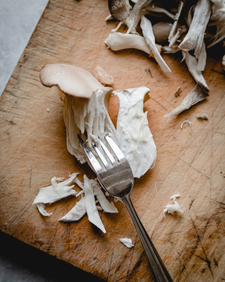 An overhead shot shows a fork "pulling" shreds of a king oyster mushroom on a wooden cutting board.