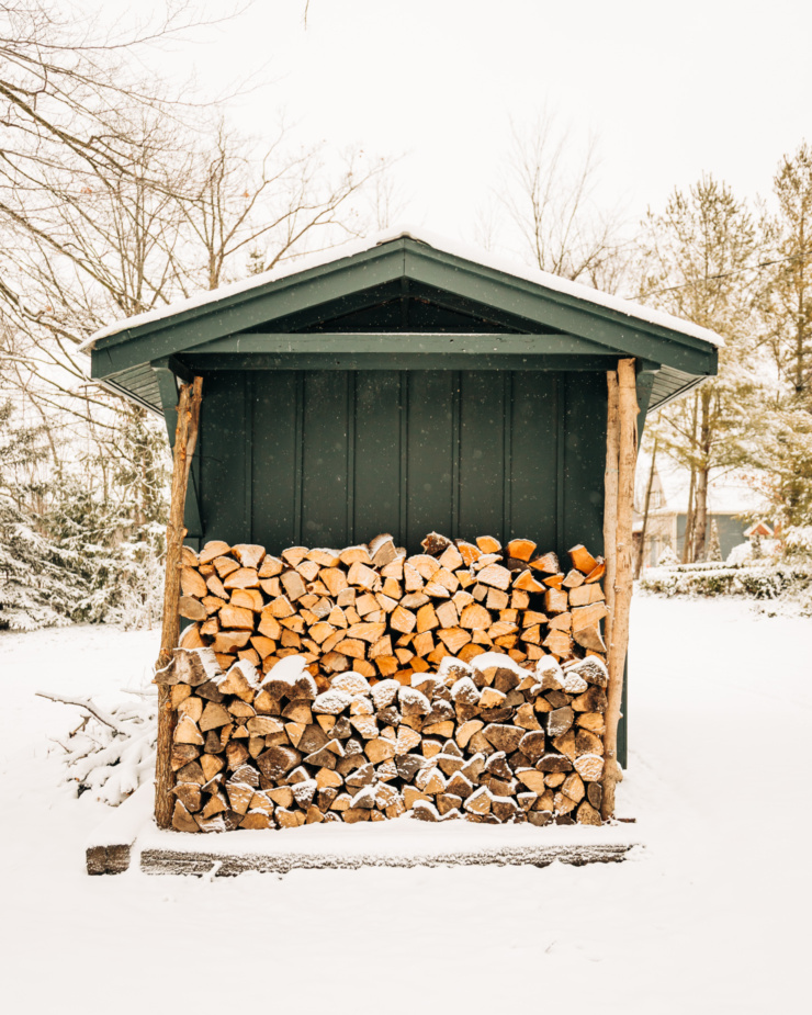 A head-on shot shows piles of chopped wood against the outside wall of a dark green outdoor shed. There are trees in the background and everything has a light dusting of snow.