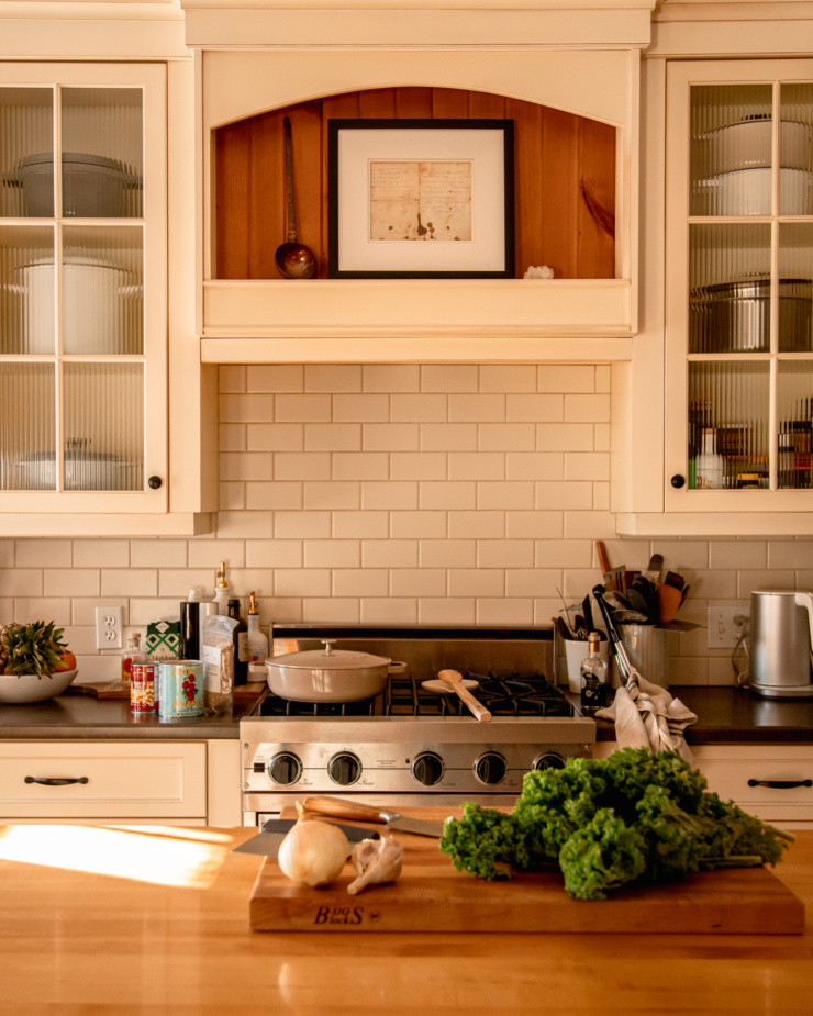 A head-on shot of a kitchen scene, featuring a pot on the stove, and some kale, onion and garlic on a chopping block.