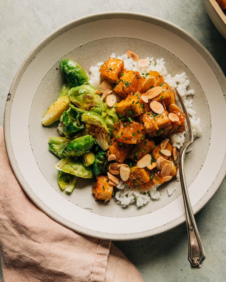 An overhead shot shows a portion of sticky marmalade tofu over some white rice with a side of seared Brussels sprouts. It is plated in a shallow bowl with a pink linen napkin to the side.
