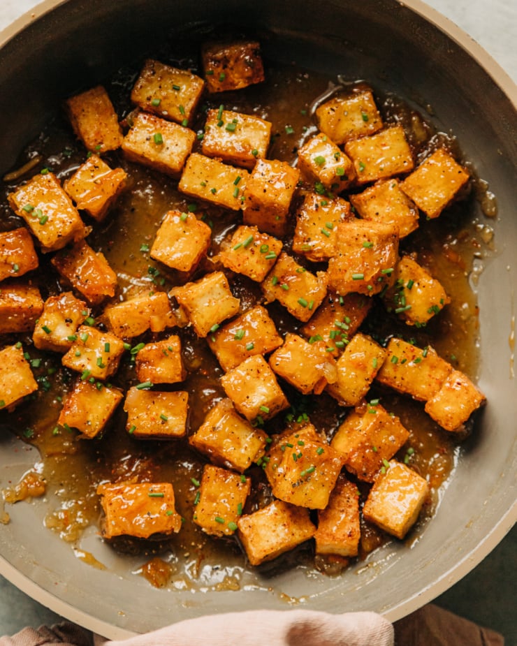 An overhead shot shows sticky marmalade tofu in a non-stick skillet. The tofu cubes are swimming in a bit of the marmalade glaze and is garnished with chopped chives. A pink linen is bunched around the skillet's handle.