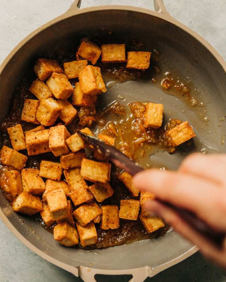 A hand is using a wooden utensil to stir crisp tofu cubes in a marmalade glaze in a non-stick skillet.