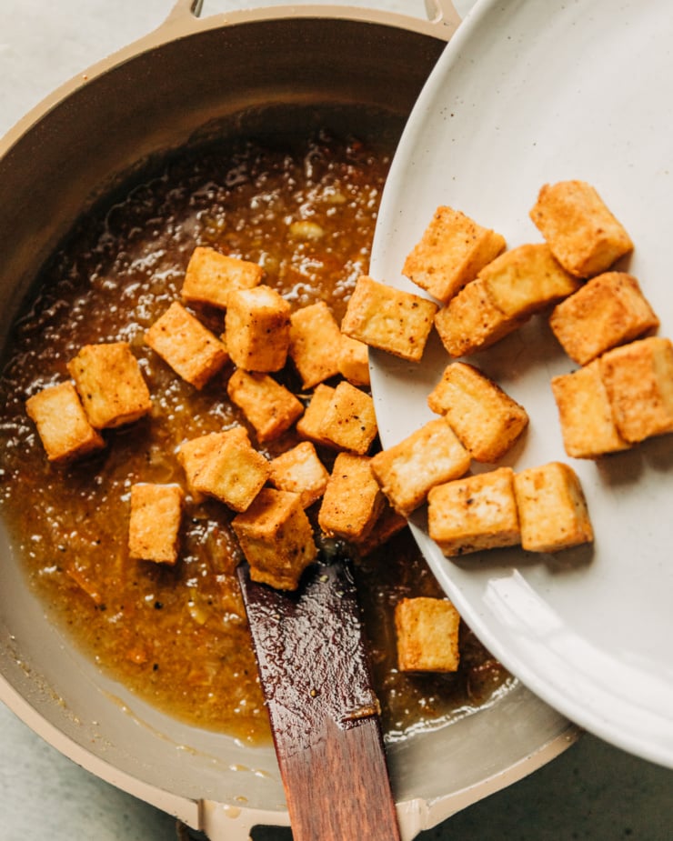 Crispy tofu cubes are being transferred from a plate to a nonstick skillet with slightly reduced marmalade glaze.