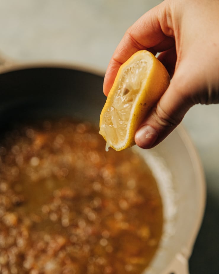 A hand is squeezing a lemon into a spiced marmalade glaze in a non-stick skillet.