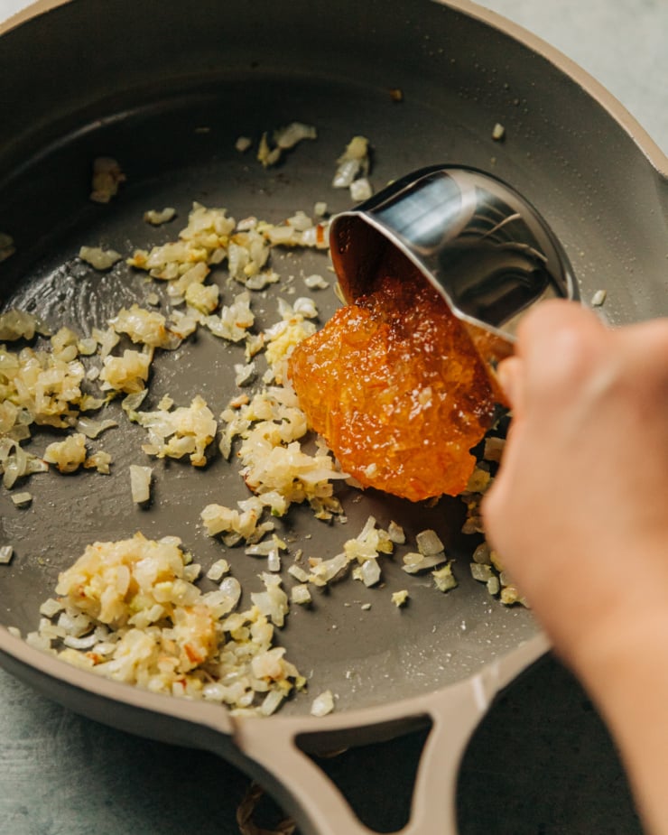 A hand is transferring orange marmalade to a skillet with sautéed onions, garlic, and ginger.