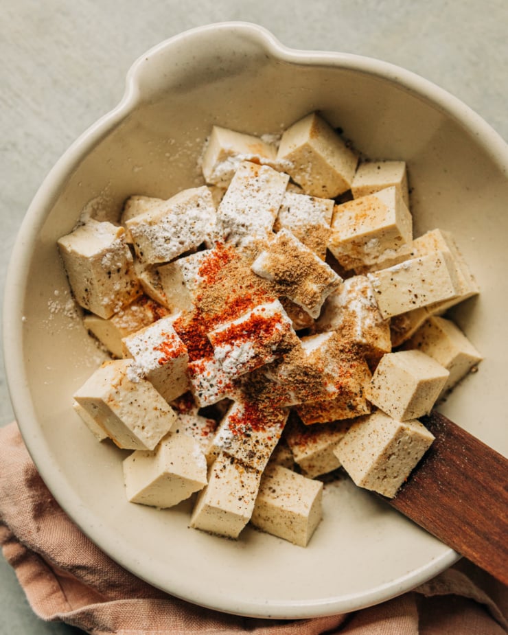 An overhead shot shows tofu cubes covered with spices and arrowroot starch. all in a small bowl with a wooden utensil for stirring.