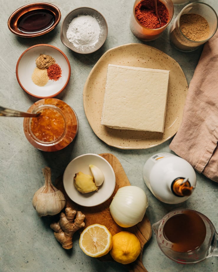 An overhead shot shows the ingredients used for sticky marmalade tofu: pressed tofu, garlic, ginger, spices, orange marmalade, lemon, olive oil, vegetable stock, Tamari, and arrowroot starch.
