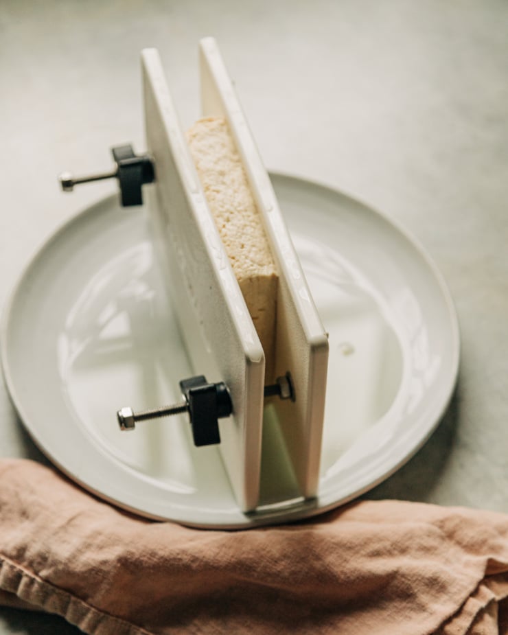 A 3/4 angle shot shows tofu being pressed over a plate,