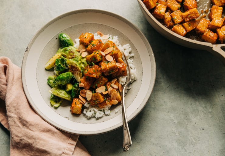 An overhead shot shows a portion of sticky marmalade tofu over some white rice with a side of seared Brussels sprouts. It is plated in a shallow bowl with a pink linen napkin to the side.