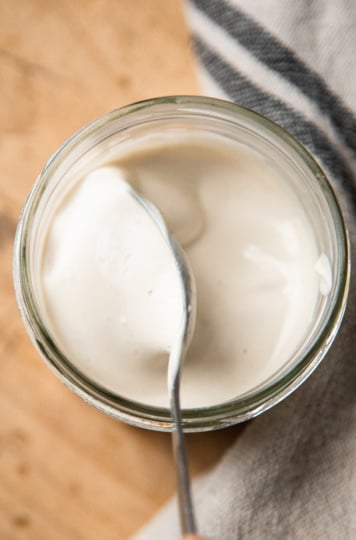 An overhead shot shows a spoon stirring up some vegan sour cream in a glass jar.