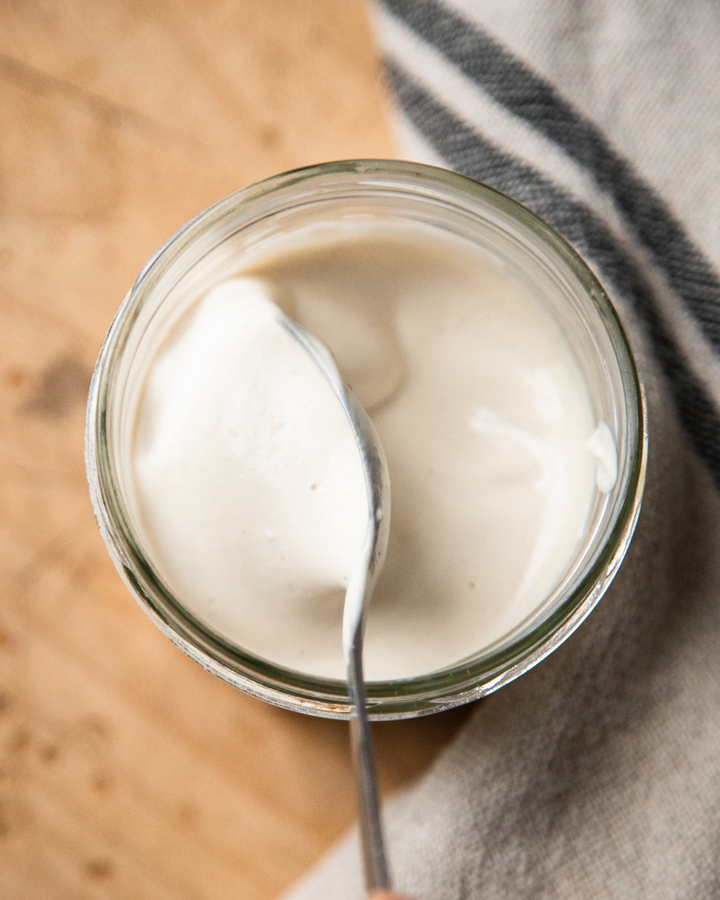 An overhead shot shows a spoon stirring up some vegan sour cream in a glass jar.