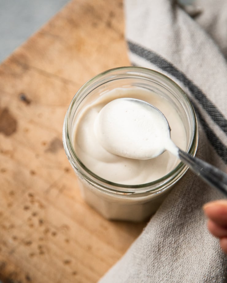 An overhead shot shows a spoon dipping into a jar of vegan sour cream. A beige linen dish towel is nearby.
