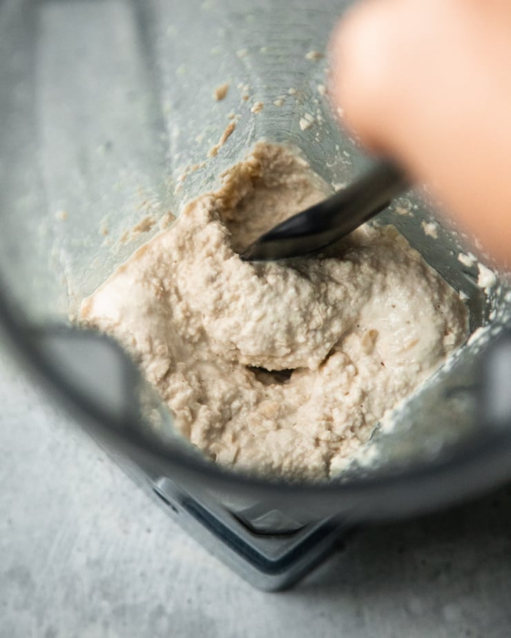 An overhead shot shows a hand using a spatula to scrape the sides down of a blender. A chunky puréed cashew mixture is in the blender.