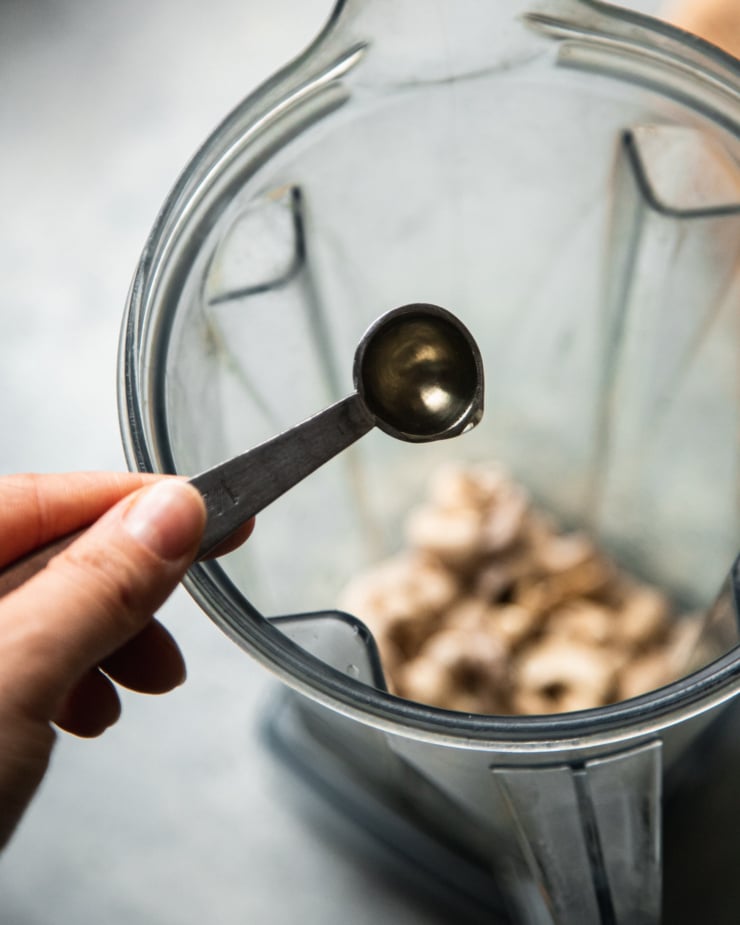 An overhead shot of a hand using a teaspoon measure to add some apple cider vinegar to a blender pitcher.