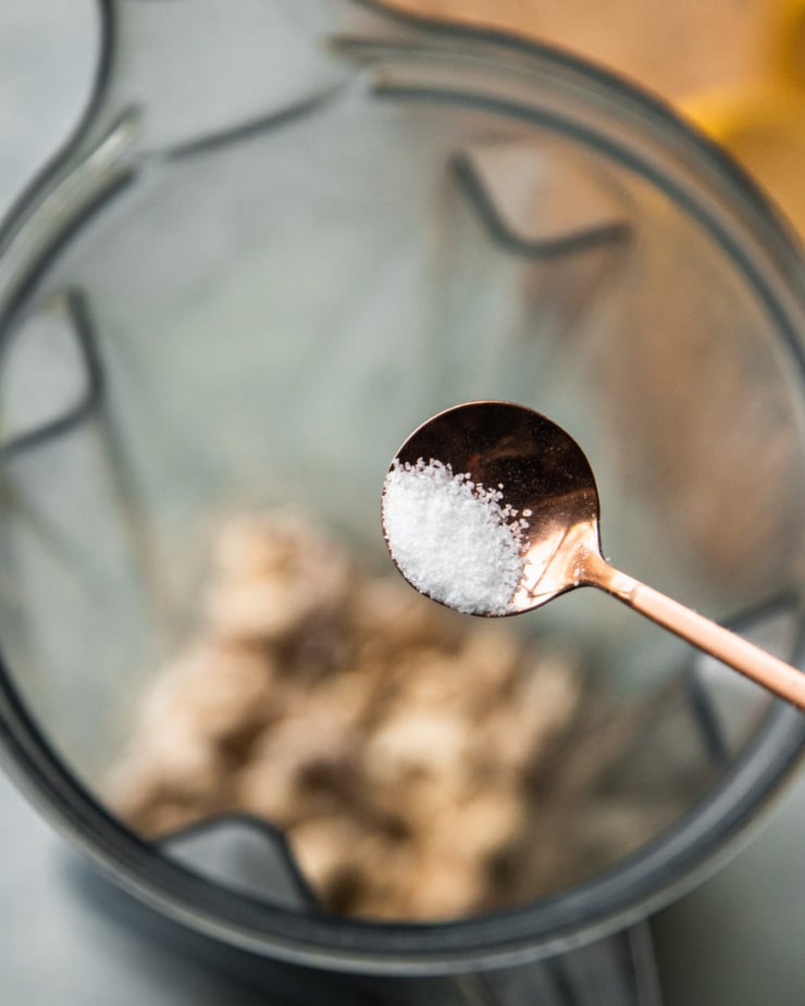 An overhead shot of a spoonful of salt being held over a blender pitcher, about to be added in.