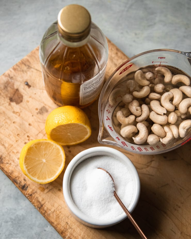 An overhead shot of ingredients used in a tangy vegan sour cream: apple cider vinegar, raw cashews that have been soaked, salt, and lemon.
