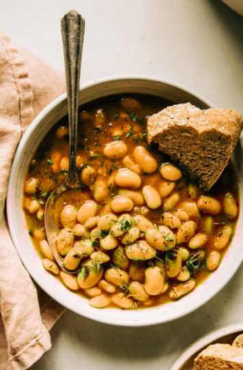 An overhead shot of an individual serving of brothy beans in a bowl. The beans are topped with chopped herbs and there is a piece of bread being dipped into the broth.