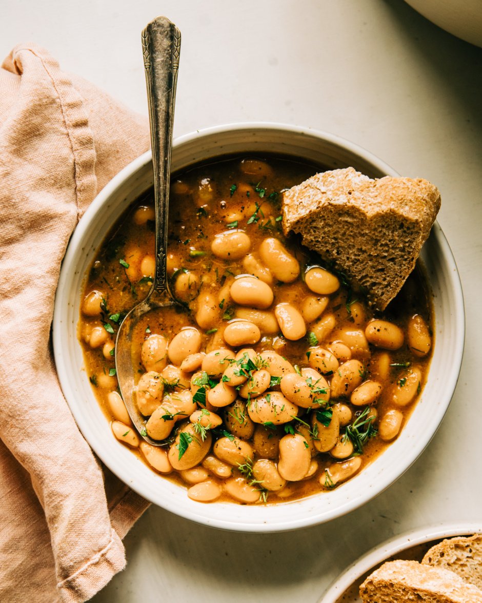 An overhead shot of an individual serving of brothy beans in a bowl. The beans are topped with chopped herbs and there is a piece of bread being dipped into the broth.