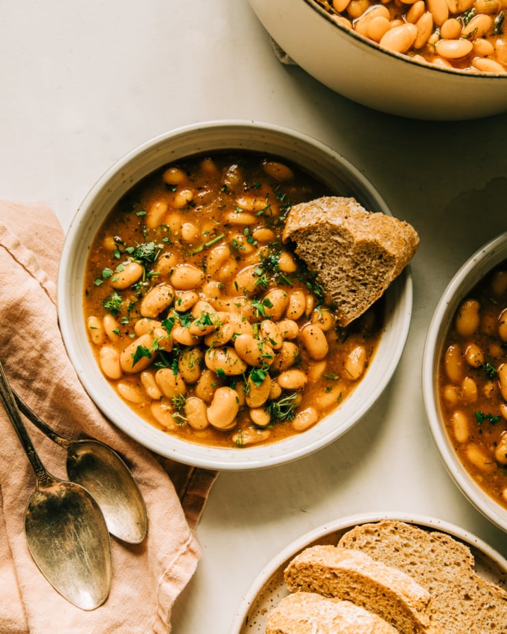 An overhead shot of an individual serving of brothy beans in a bowl. The beans are topped with chopped herbs and there is a piece of bread being dipped into the broth.