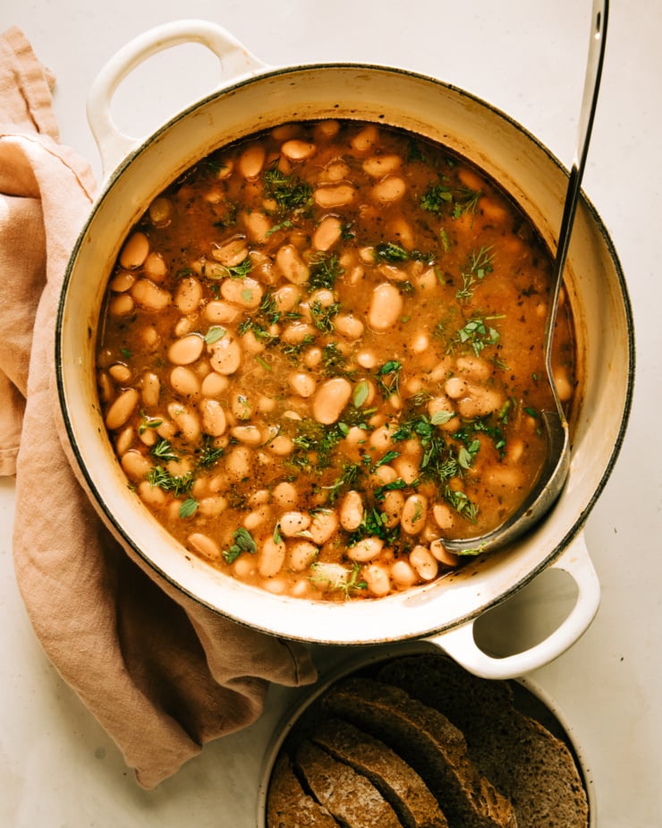 An overhead shot of a Dutch oven filled with a batch of brothy beans. The beans are garnished with chopped herbs and a ladle is sticking out of the pot. A plate with sliced bread is seen nearby.