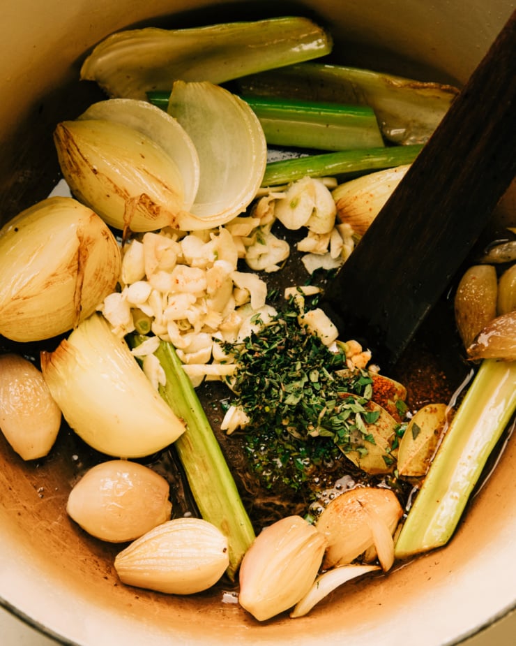 An overhead shot of onion quarters, celery sticks, shallots, garlic, herbs, and spices in a Dutch oven. There is light browning on the onions.