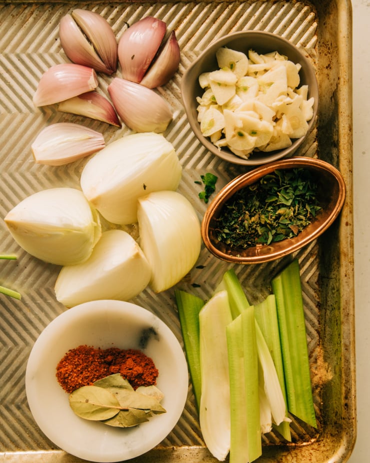 An overhead shot of ingredients that are prepped: shallots and onions are quartered through the root, celery is cut into sticks, garlic cloves are sliced, spices are measured, and herbs are minced. All photographed on an old baking sheet.