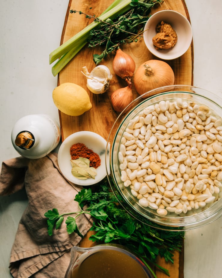 An overhead shot of raw ingredients used in a brothy beans recipe. There is: celery, thyme, oregano, miso, shallots, onions, lemon, garlic, olive oil, spices, bay leaves, dill, parsley, soaking white beans, and vegetable stock. All photographed on a wooden board.