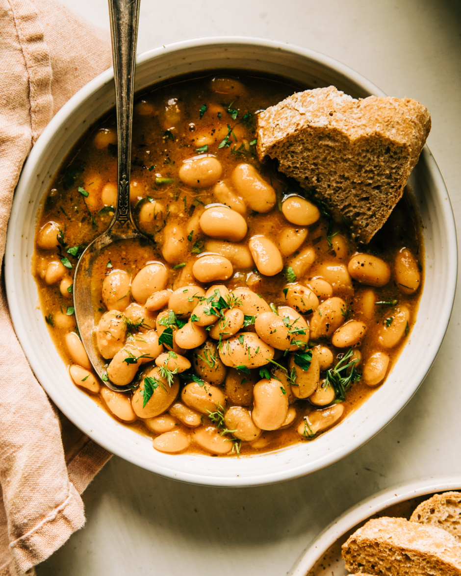 An overhead shot of an individual serving of brothy beans in a bowl. The beans are topped with chopped herbs and there is a piece of bread being dipped into the broth.