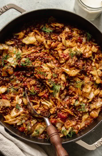 An up close, overhead shot shows a finished vegan cabbage roll skillet with french lentils, chopped tomatoes, rice, walnuts, and spices. The skillet is topped with fresh dill and a jar of vegan sour cream is seen nearby.