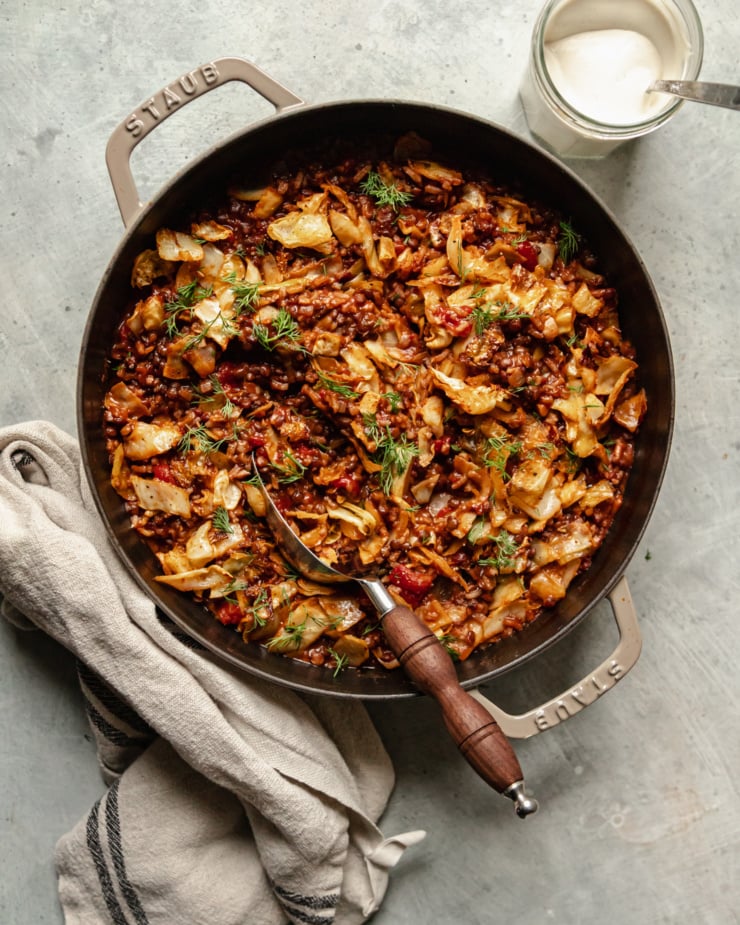 An overhead shot shows a finished vegan cabbage roll skillet with french lentils, chopped tomatoes, rice, walnuts, and spices. The skillet is topped with fresh dill and a jar of vegan sour cream is seen nearby.