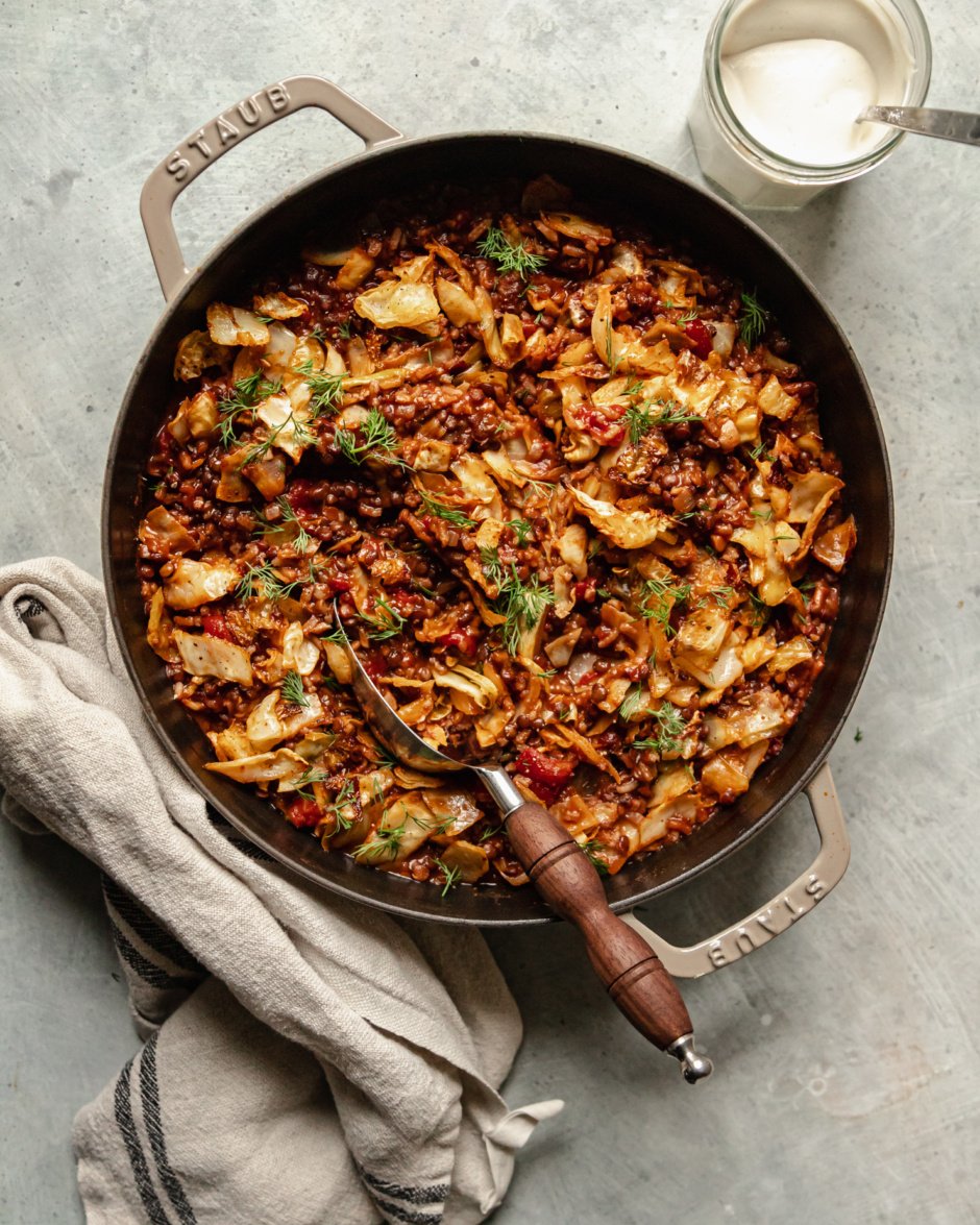 An overhead shot shows a finished vegan cabbage roll skillet with french lentils, chopped tomatoes, rice, walnuts, and spices. The skillet is topped with fresh dill and a jar of vegan sour cream is seen nearby.