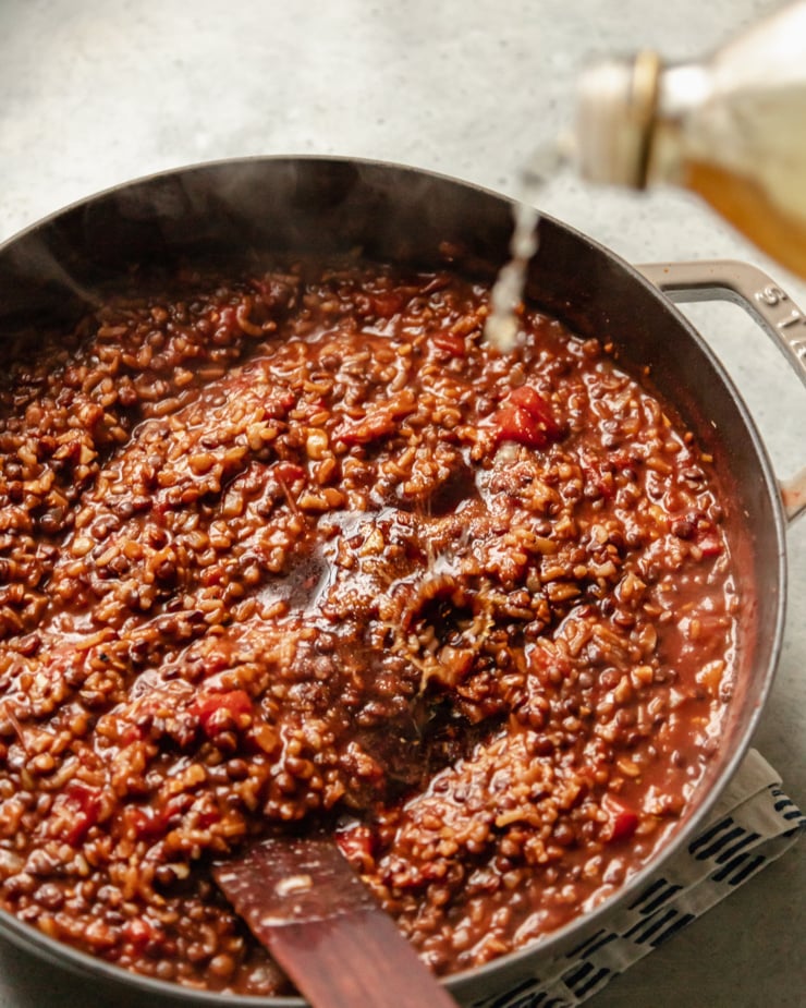 A slight 3/4 angle shot shows apple cider vinegar being poured into a pot of stewing lentils, rice, tomatoes, walnuts, onions, and spices.