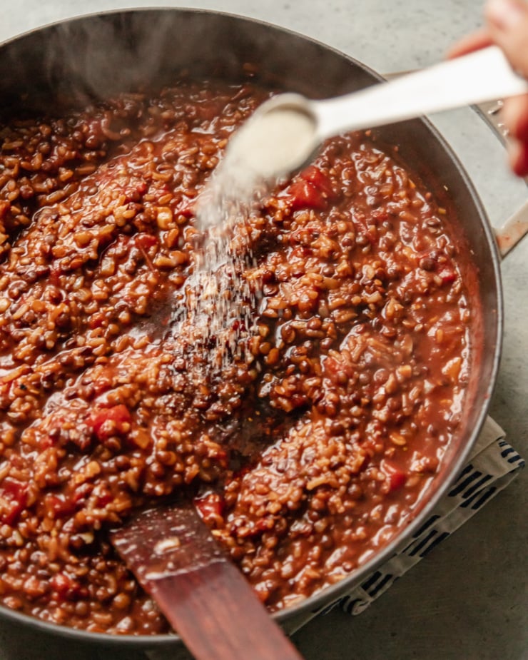 A slight 3/4 angle shot shows a spoon of sugar being sprinkled into a pot of stewing lentils, rice, tomatoes, walnuts, onions, and spices.