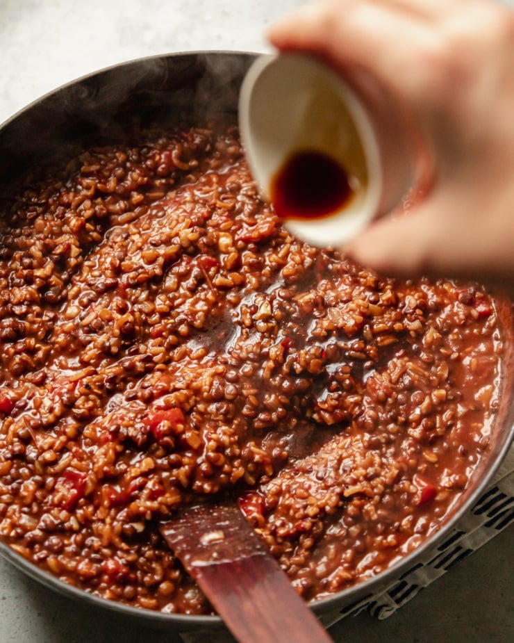 A slight 3/4 angle shot shows a hand pouring some Tamari into a pot of stewing lentils, rice, tomatoes, walnuts, onions, and spices.
