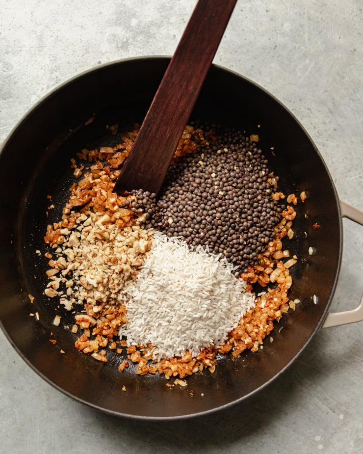 AN overhead shot shows dried lentils, white rice, and finely minced walnuts added to a pot with seasoned sautรฉed onions.