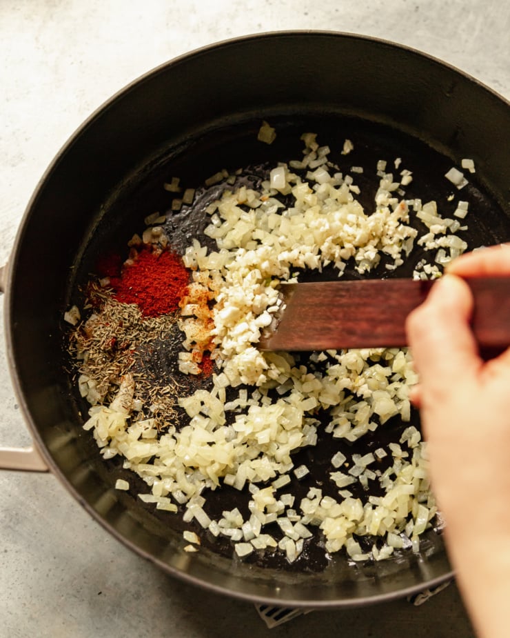 An overhead shot shows a hand using a wooden utensil to stir spices and garlic into some sautรฉed onions in a braiser-style pot.