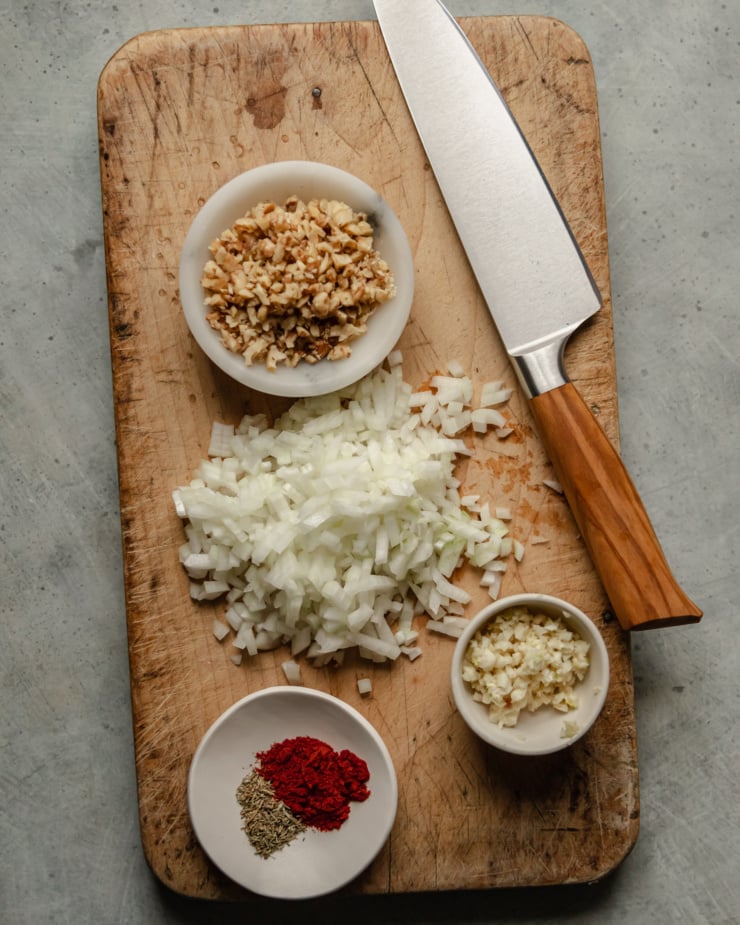 An overhead shot shows finely diced onions, walnuts, minced garlic, and measured out dried spices on a worn wooden cutting board.