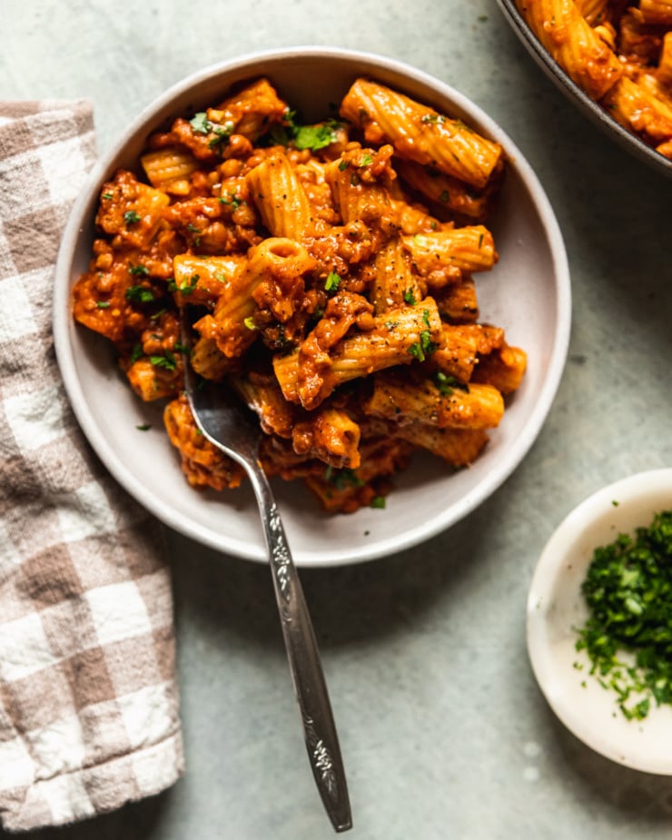 An overhead shot of a serving of lentil bolognese rigatoni in a wide, white bowl. A checkered napkin is nearby and the pasta is garnished with finely chopped parsley.